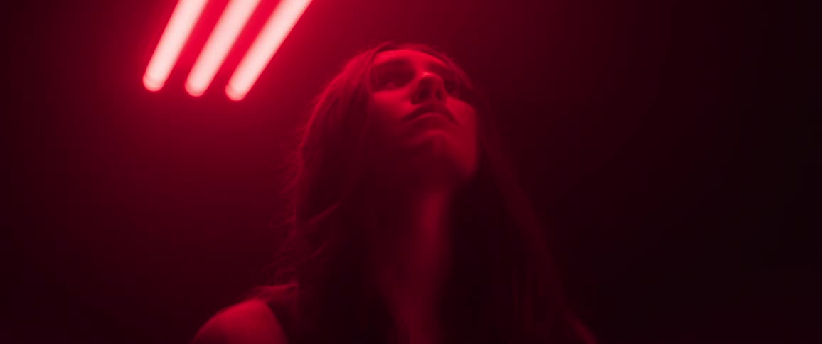 A woman with long dark hair is looking upward illuminated by red neon lights from above in a dark room