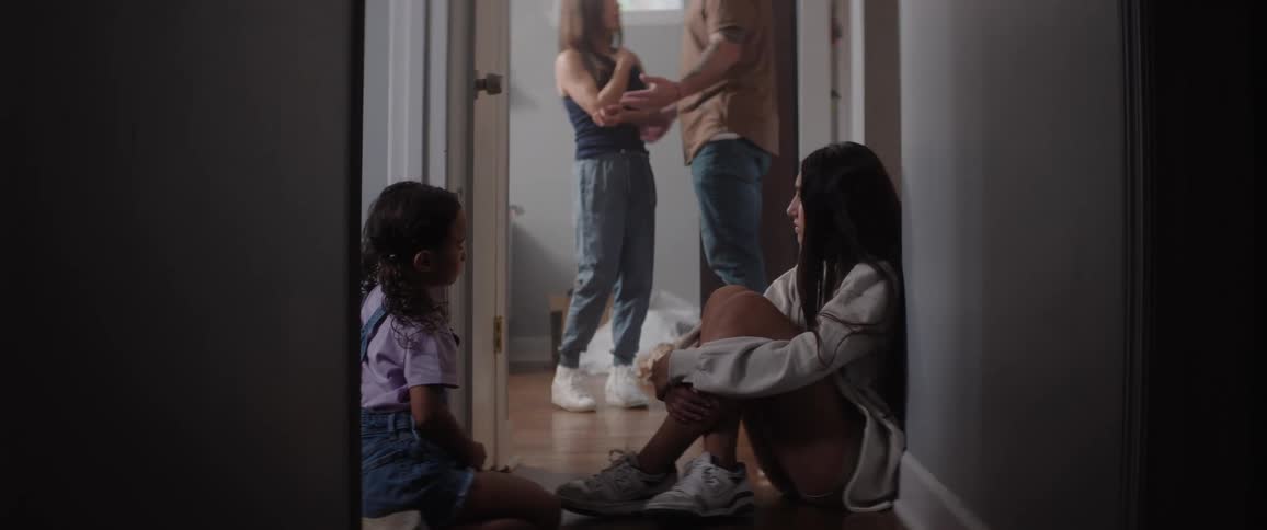 Two sad children sit in a hallway as their parents argue in the background