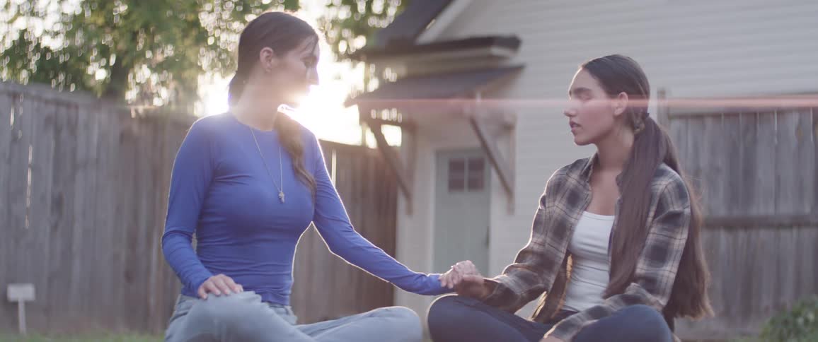Mother and daughter sit on grass facing each other with their legs crossed in a backyard setting with a house and fence visible in the background