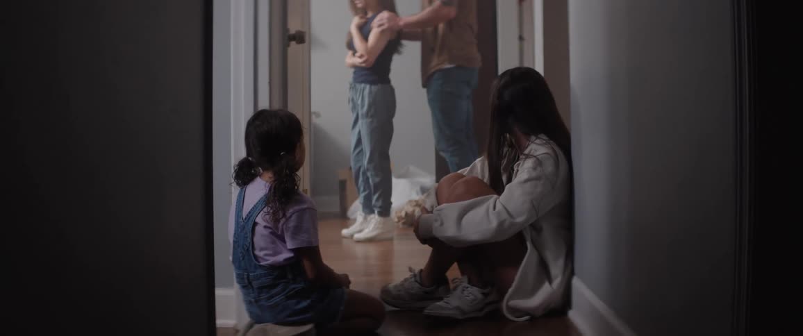 Daughters sit in the hallway and watch their parents talk with concern in a bedroom