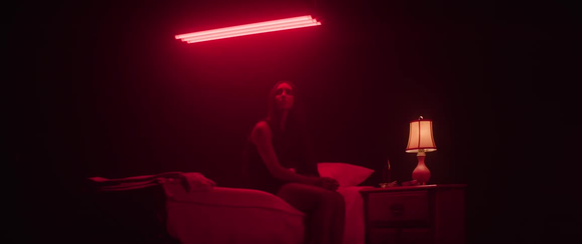 A woman sits on a bed under red lighting in a dark room with a dimly lit nightstand