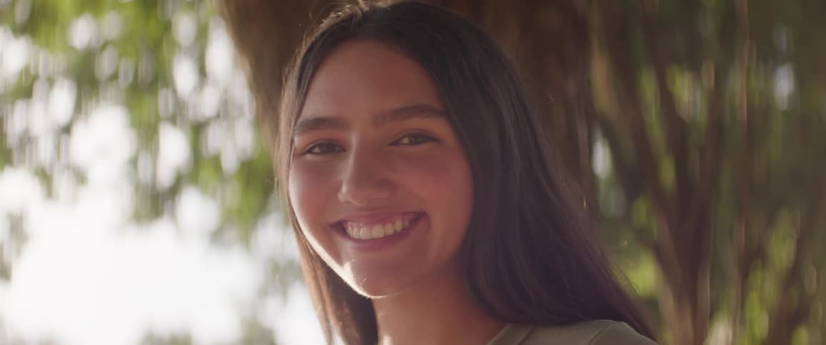 The face of a girl like teenager who is smiling looking straight ahead in an outdoor environment with a tree in the background