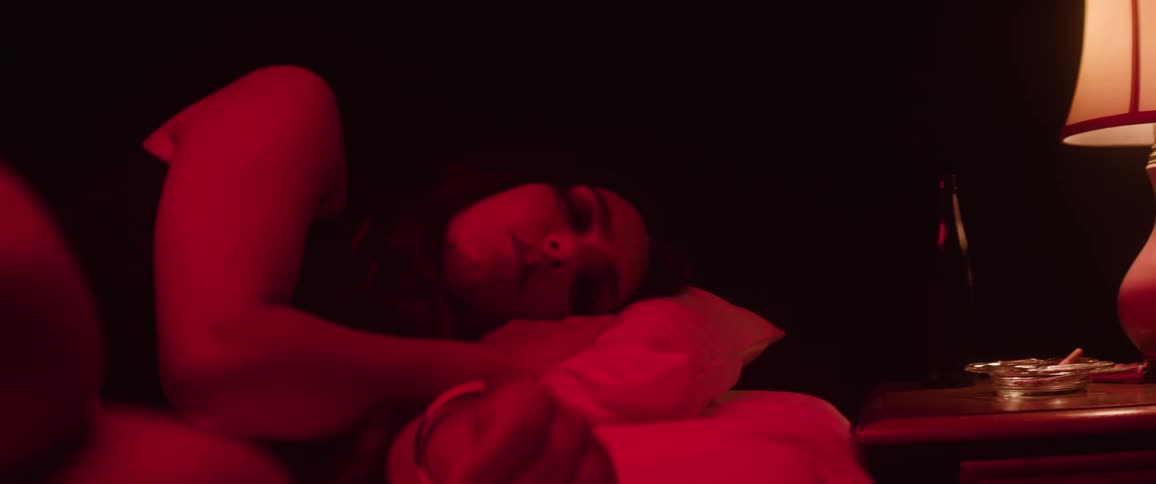 A woman lies in a bed with red lighting next to a nightstand with a lit lamp and an ashtray