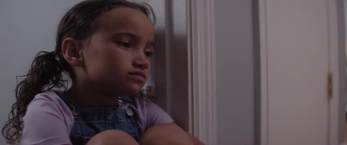 A little girl sits alone in a room against a wall looking very sad