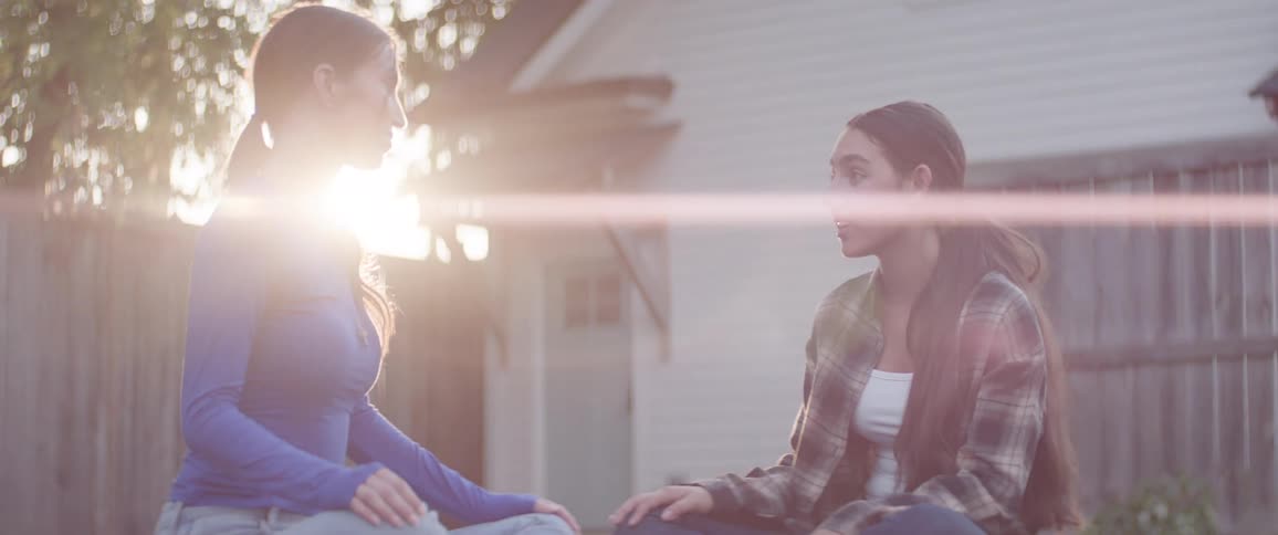 Two teenage girls are sitting on the floor outside talking and laughing while the sun sets behind them