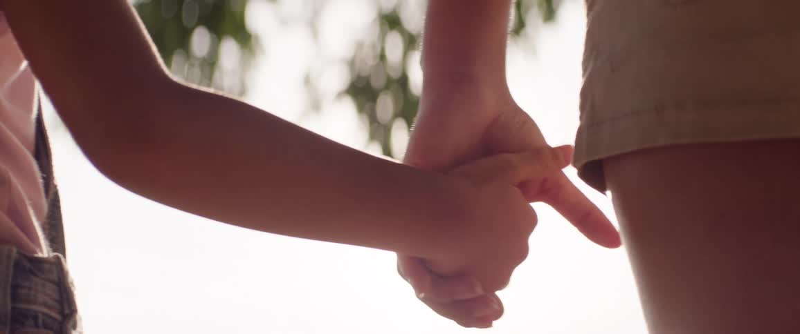 An adult and a child hold hands against a bright background with sunlight