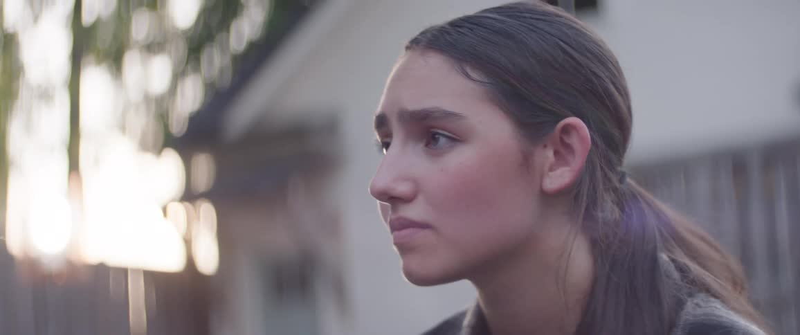 Woman with long dark hair in a ponytail gazes rightward with a sad expression a fence and bright background behind her