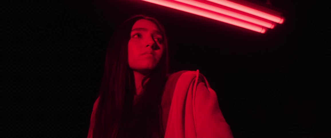 A woman stands under red fluorescent lighting in a dark setting wearing a red outfit