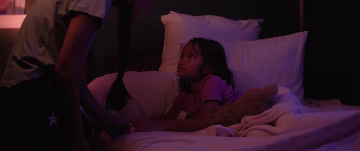 A scared child smiles from bed while making a pinky promise with a woman in a dimly lit room bathed in purple ambient light