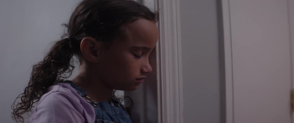 A ittle girl whos leaning against a doorframe and shes looking down in sad tones