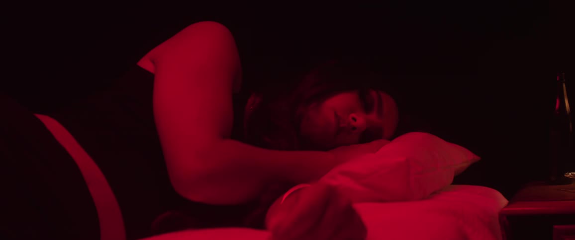 Beautiful woman lies on a bed under red lighting beside a nightstand holding a bottle and other small items