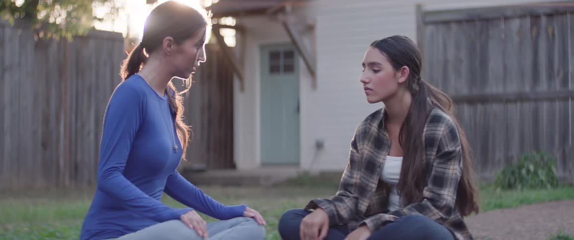 Two young women sit outdoors on the sidewalk during the daytime talking