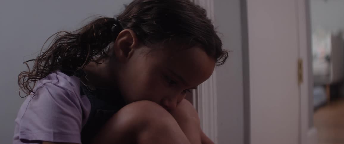 A young girl sits curled up against a wall appearing sad and withdrawn