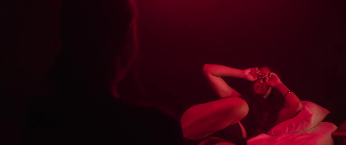 A woman lying on a student bed in a dark red room is awakened by another woman