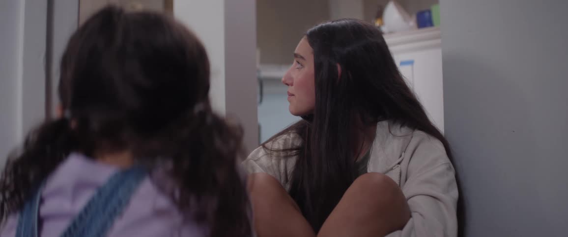 A teenager sits on the floor indoors looking at a child before turning her face to the left with a sad expression