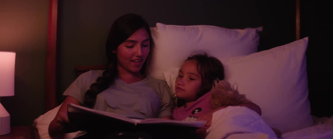 Teenage sister reads a book aloud while her younger sister listens holding a stuffed animal as they lie together in bed