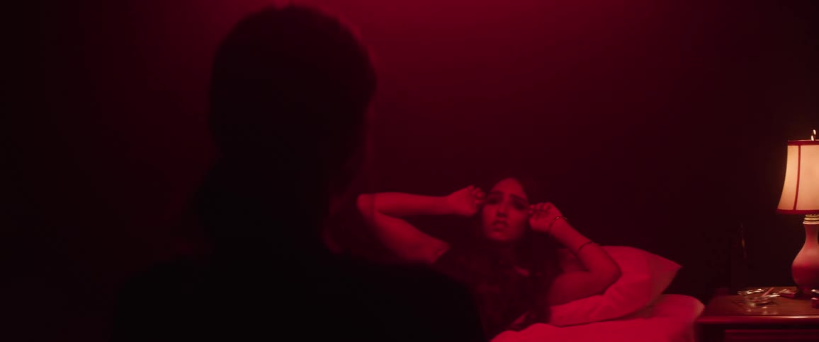 A silhouetted figure faces a reclining person who stirs awake startled and dazed as deep red light envelops them in a dreamlike atmosphere