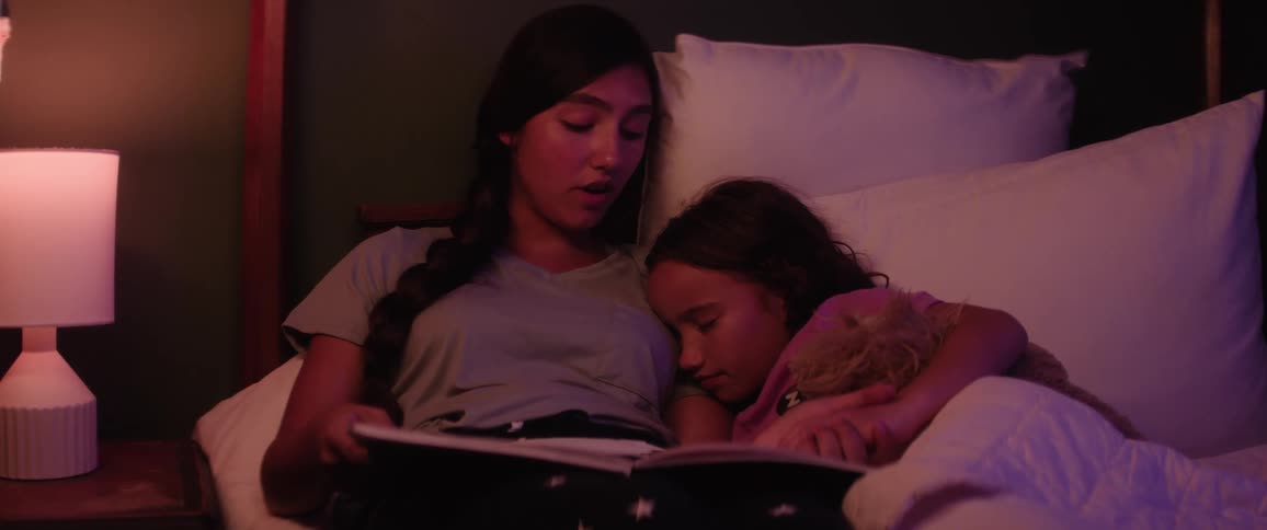 Female teenager reads to her younger sister in bed to sleep in a room at night