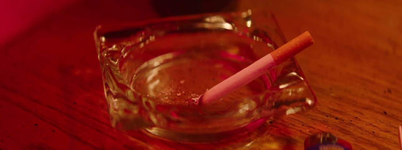 Close up of a burning cigarette in a glass ashtray placed on a table inside a red lit room