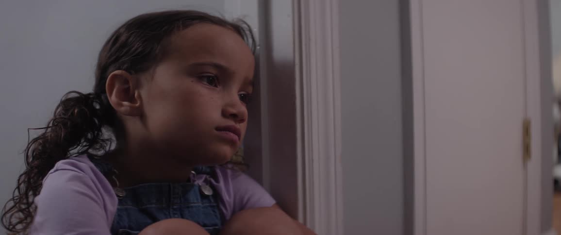 A sad child in purple and denim overalls sits on the floor with knees tucked leaning back against a white wall