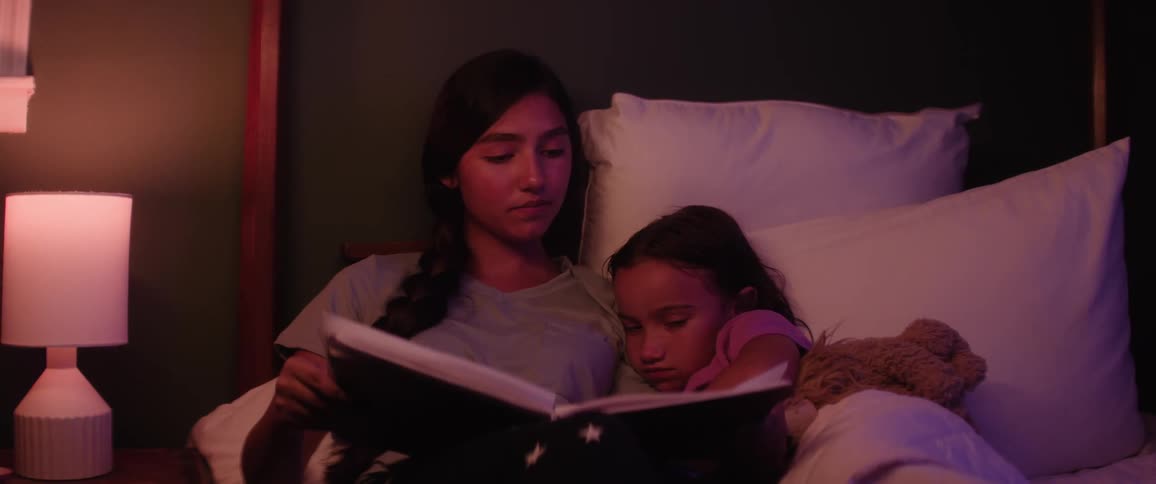 A teenage girl reads a book to her younger sister in bed at night before bedtime
