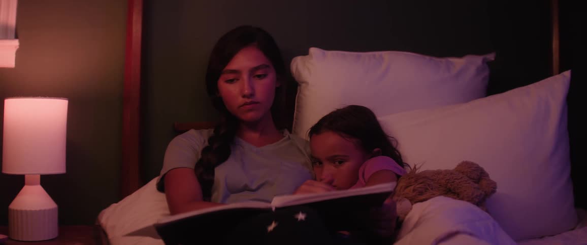 Female teenager reads a book to her sister in bed at night before bedtime