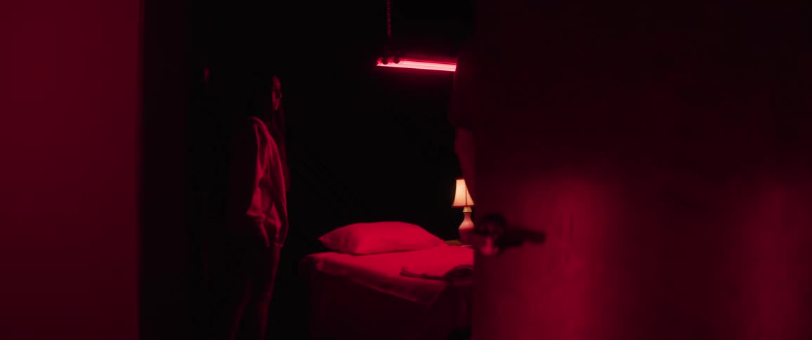 Two people stand near a bed in a dimly lit red room as the one closest to the door closes it
