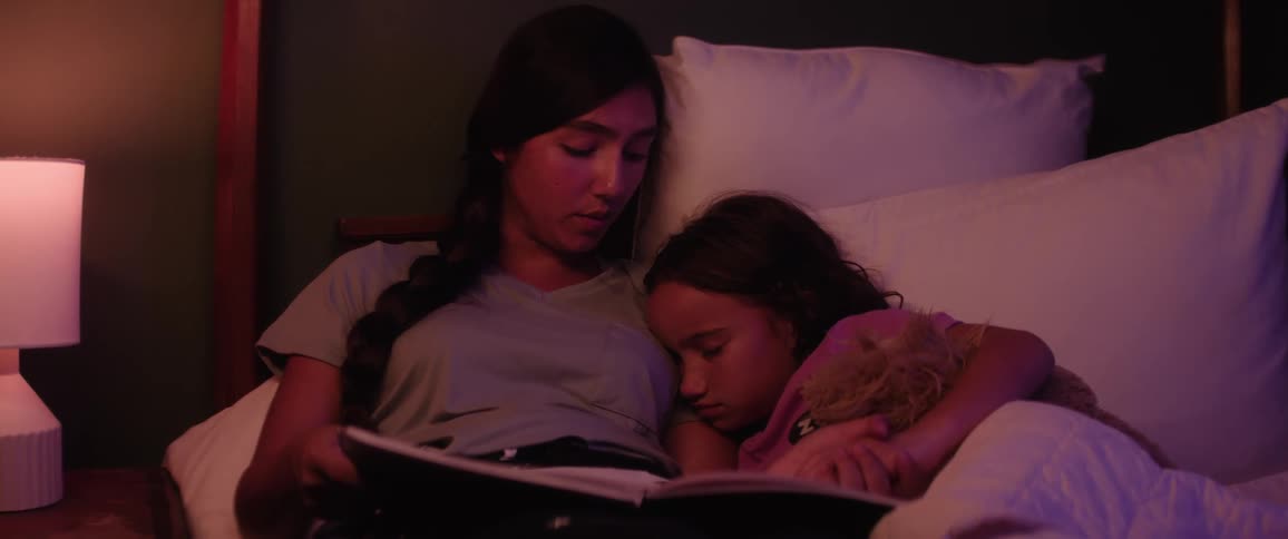 An older girl reads a bedtime story to her younger sister who is resting beside her