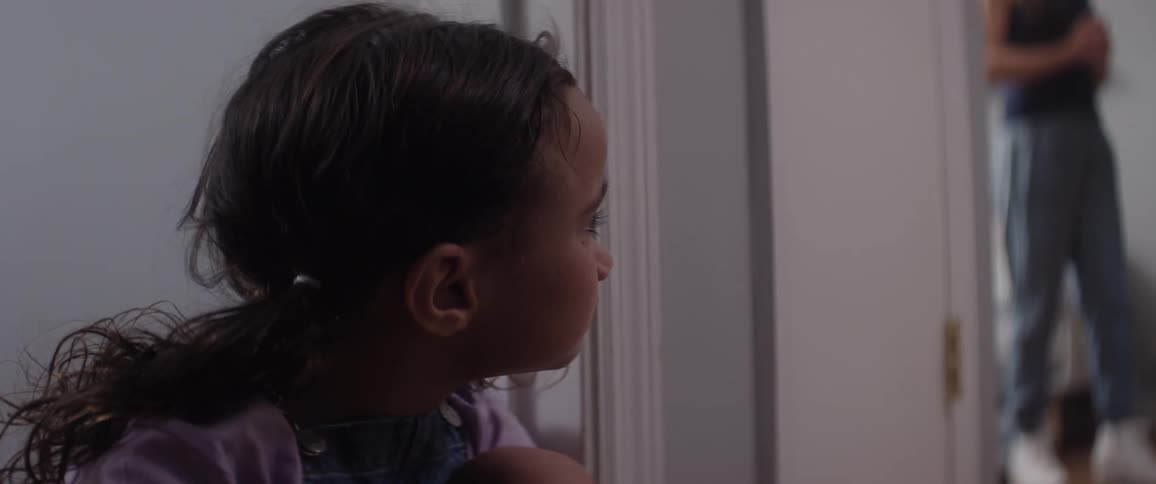 A girl is sitting next to a door and she is sad as she is looking at an adult person standing in another room