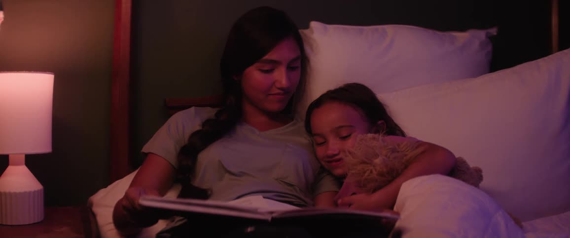 An older sister reads a book to her younger sister in bed under warm ambient light