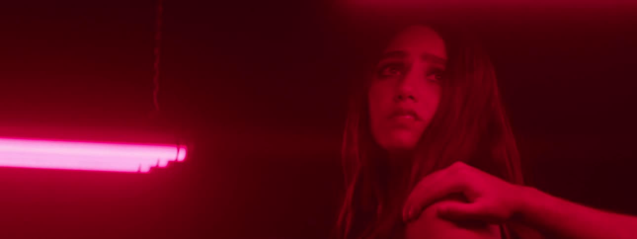 A woman stands in a dark space illuminated by red light as someone grips her shoulder