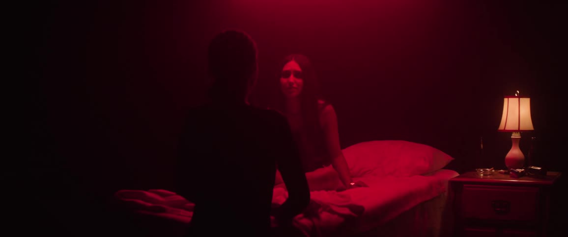 A person sits on a bed in a dark room illuminated by red light facing another person who is silhouetted