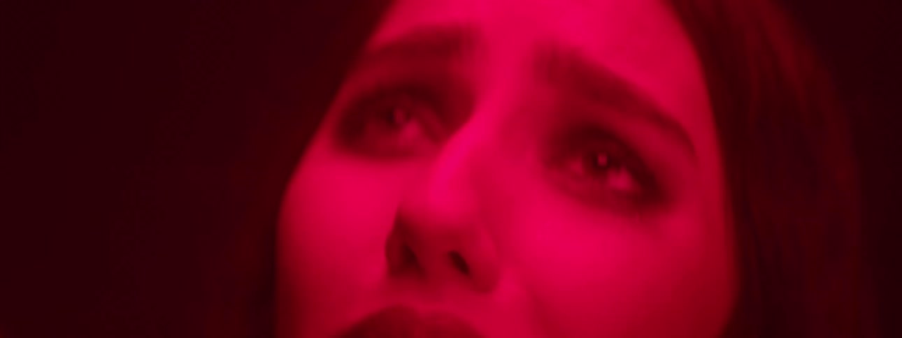 A womans face with black makeup around her eyes and she is looking up crying and a red light coloring her skin
