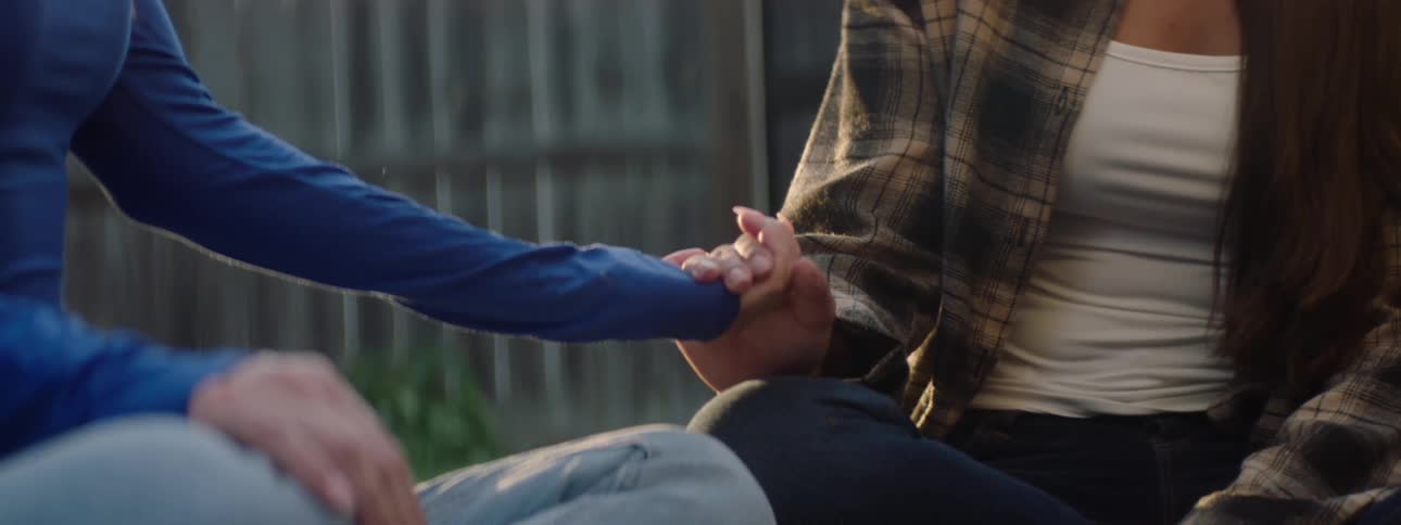 Two young women hold hands in a backyard one wearing a blue sleeve and the other a plaid shirt over a white top under morning or sunset light