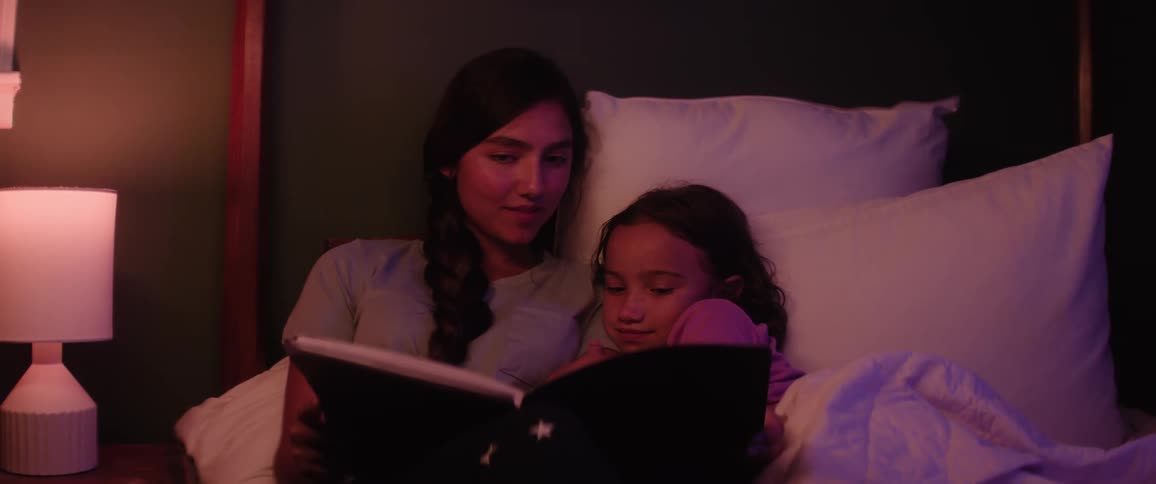 Female teenager opens and reads a book to her younger sister in bed at night before bedtime