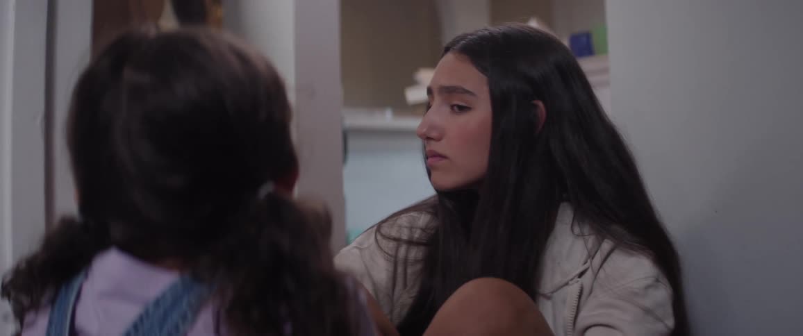 A teenager sits beside her younger sibling in the hallway gently offering comfort as they quietly observe others talking in the adjacent room