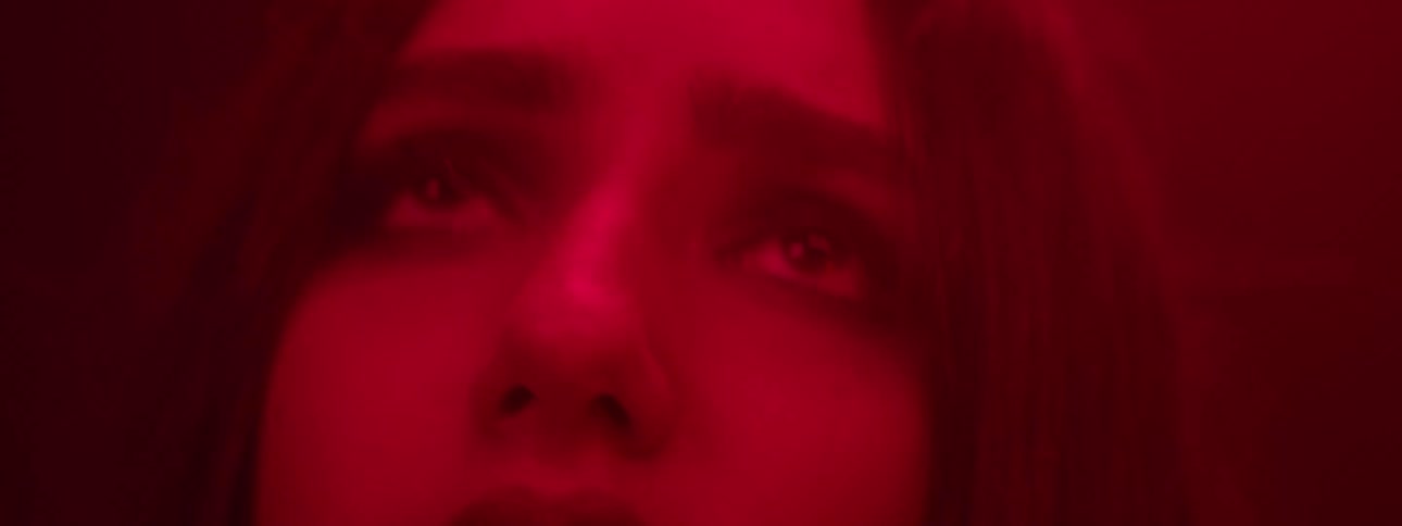 A womans face is shown in a close view bathed in red light opening her eyes and looking up