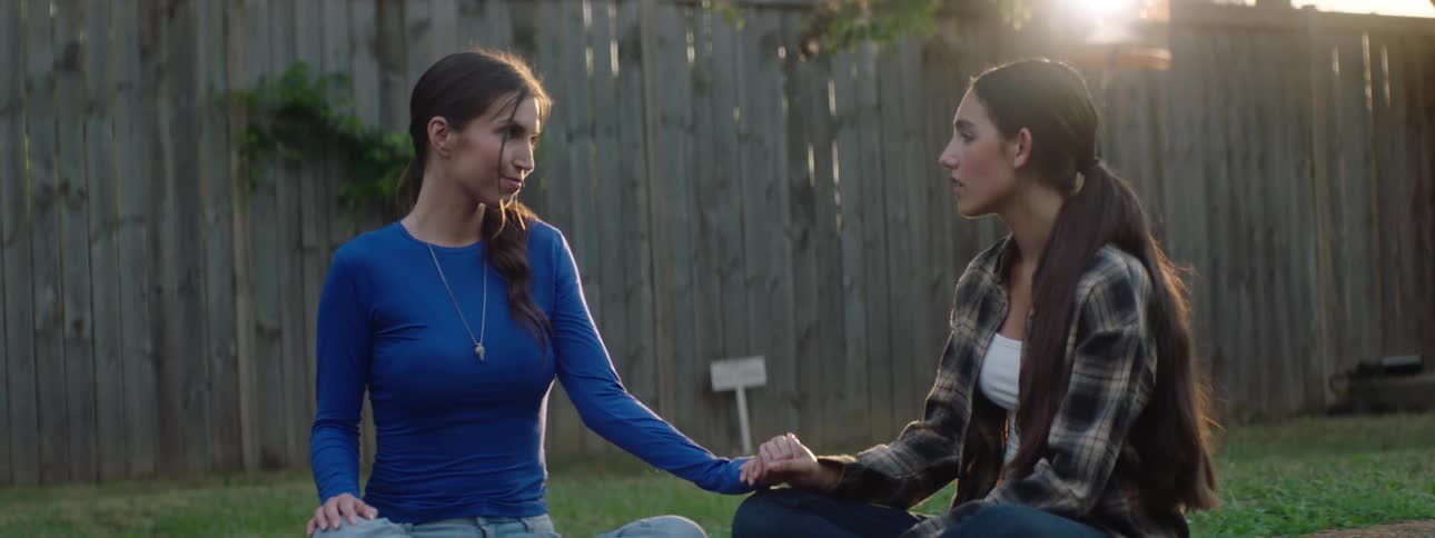 Two women sit in a backyard holding hands and sharing a quiet emotional moment together