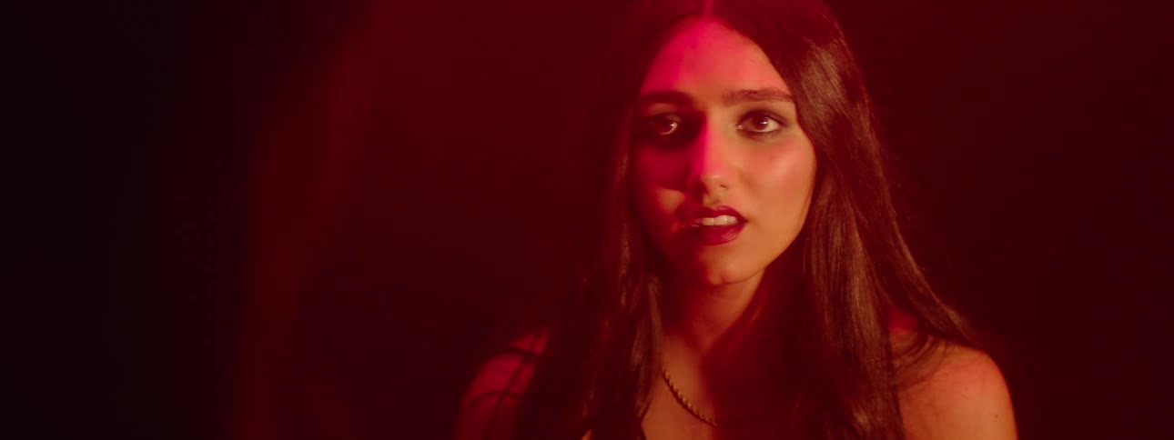 A young woman looking afraid stands facing another woman in a room with red ambient lighting