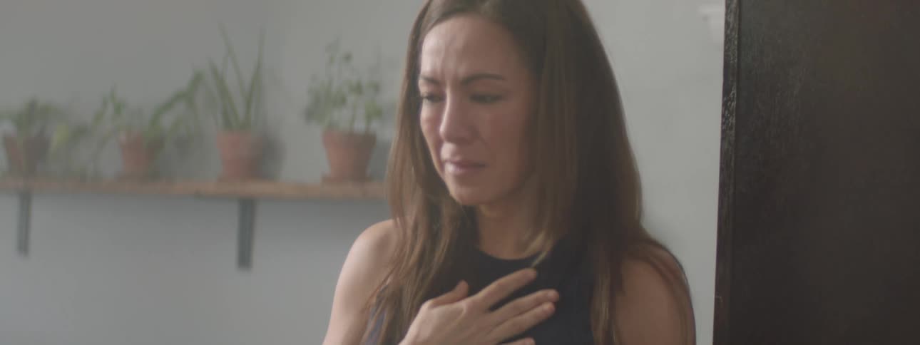 A sad woman with long brown hair holds a hand to their chest walking around a room