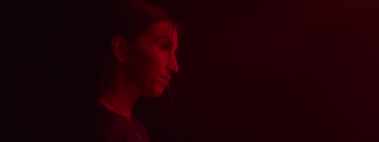 Person stands in darkness head bowed slightly as red light highlights strands of hair across their face