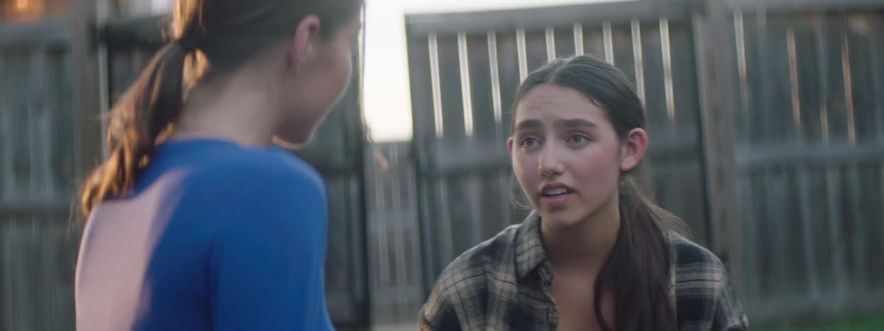 Two teenagers talk to each other outdoors near a fence at midday