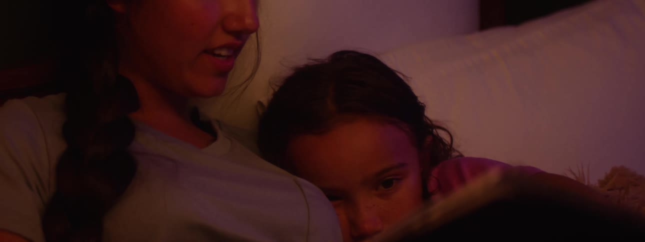 A child with dark hair lies beside a woman who is reading book that is held open in front of them in a dimly lit setting
