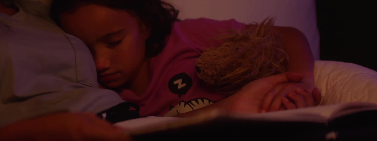 A young girl reads a bedtime story to a sleeping child as they both lie on the bed