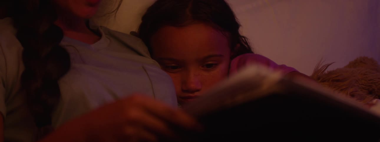 Teenage girl reads a book to her younger sister in bed at night before bedtime