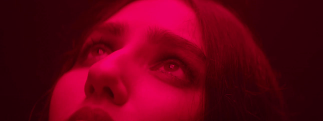 A woman face is shown in a close view bathed in red light with their eyes looking upward