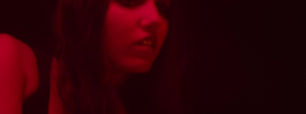 A woman with long dark hair lies down under red lighting wearing a metallic bracelet appearing confused as she wakes