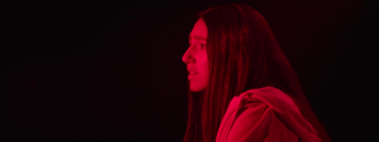A woman in a dark room illuminated by red light looks afraid as she glances around the environment
