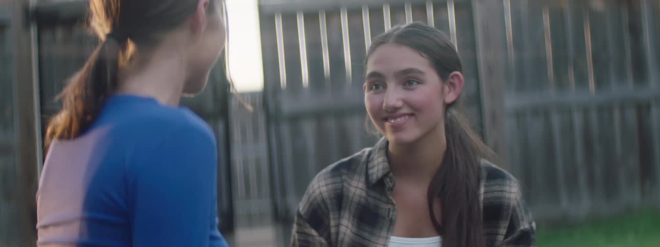 Two female teens smile and converse while standing outdoors during daytime