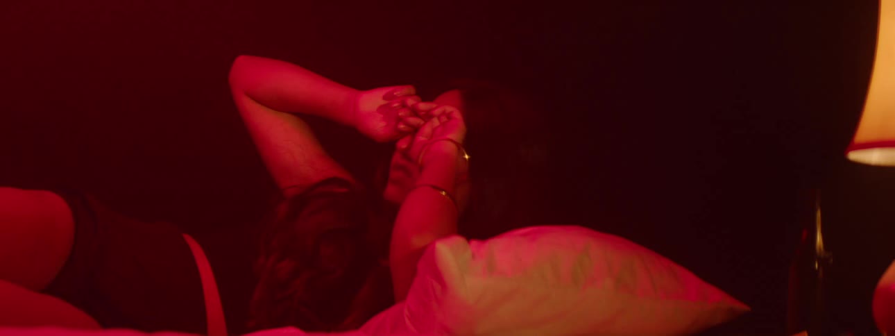A young woman wakes up in a dark room with a lamp beside her illuminated by red light looking scared